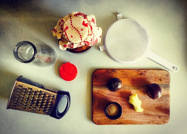Kombucha flavoring setup: a jar of kombucha covered with a cloth, a sieve, a grater, bottles, passion fruit, ginger and sugar