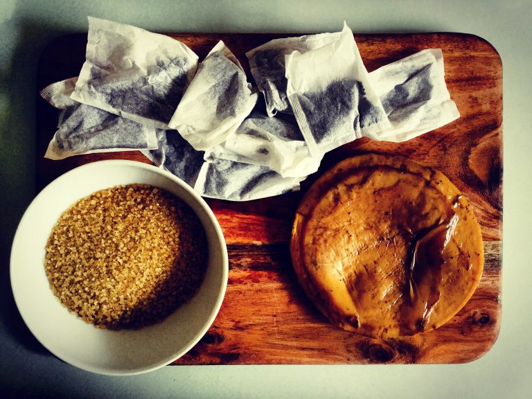Kombucha ingredients: a bowl of raw cane sugar, tea bags and a SCOBY on a wooden board
