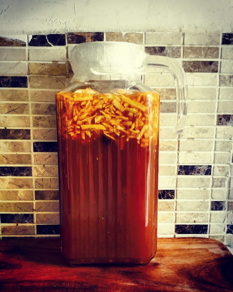 A jug of kombucha with grated apple and ginger floating on top, covered with cling film
