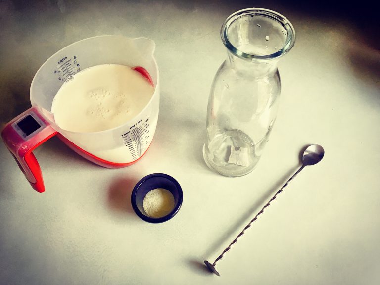Kefir-making setup: a measuring jug of milk on a scale, a small dish of powdered kefir starter, a glass bottle and a long spoon